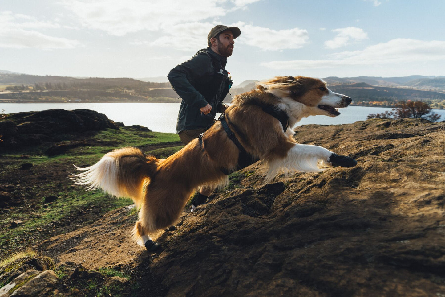 Man and dog running on a rocky hillside with a scenic landscape in the background wearing ruffwear ridgeline harness, lead and collar