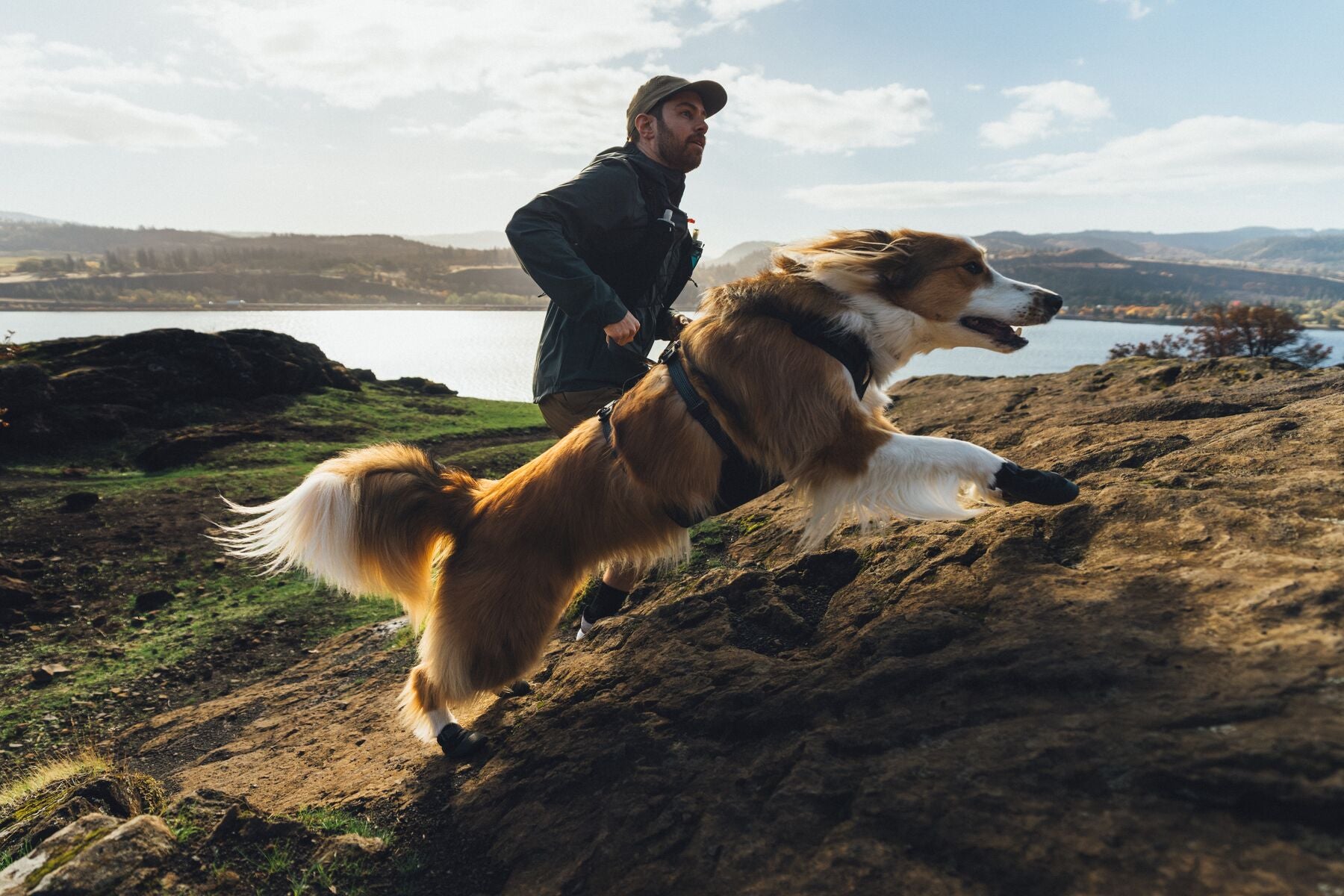 Man and dog running on a rocky hillside with a scenic landscape in the background wearing ruffwear ridgeline harness, lead and collar